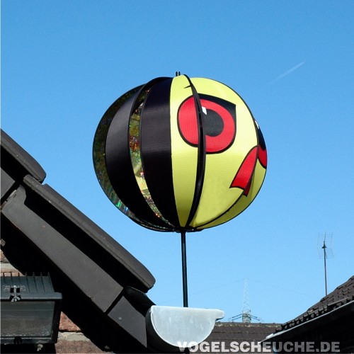Windrotor TURBINE in Gelb-Rot auf einer Dachkante, Turbinenlamellen mit Habichtaugen-Aufdruck vor blauem Himmel.